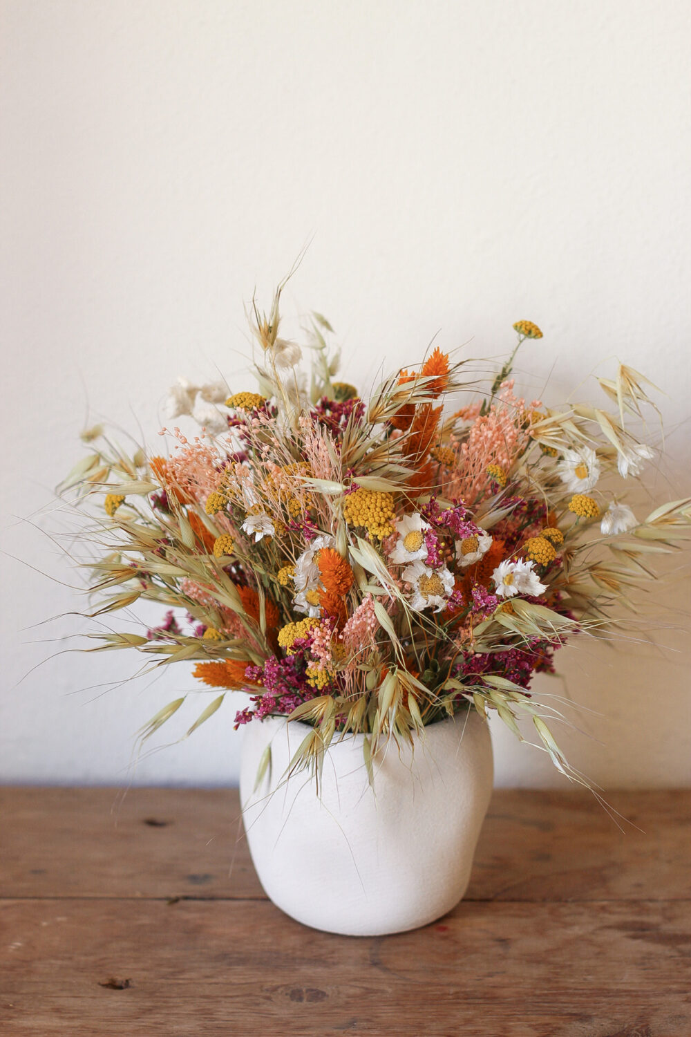 Arreglo de flores secas en tonos cálidos en jarrón blanco sobre mesa de madera rústica