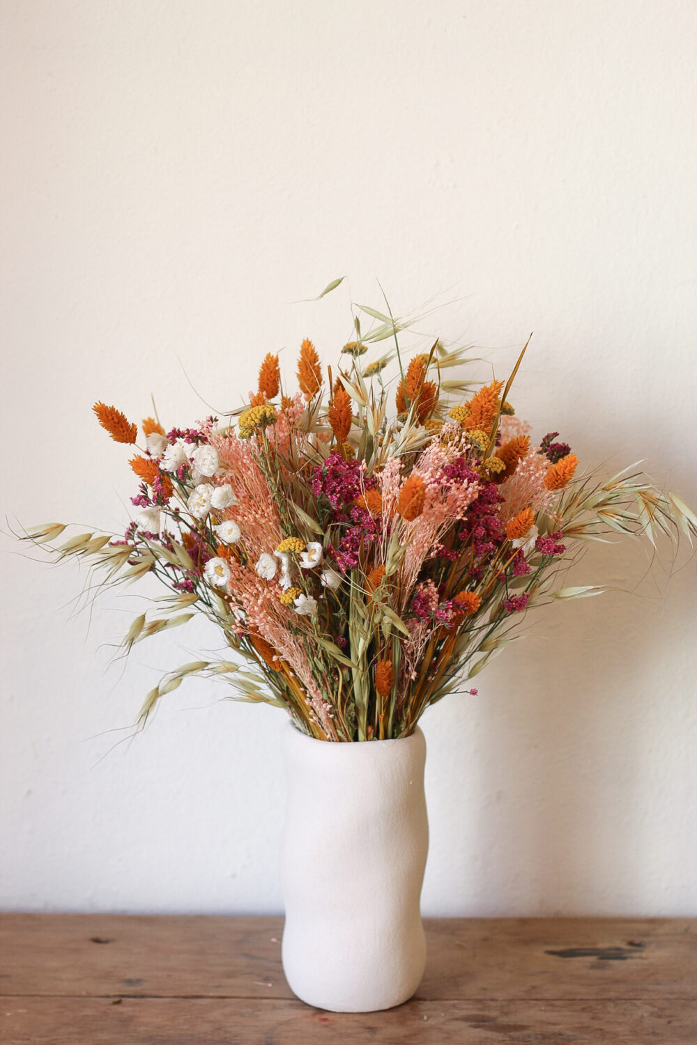 Ramo de flores secas en colores cálidos en jarrón blanco sobre mesa de madera